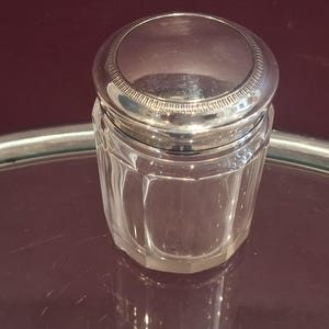 May include: A clear glass jar with a silver lid. The jar has a faceted design, and the lid has a decorative, ridged edge. The jar is sitting on a reflective surface, with a dark red background.