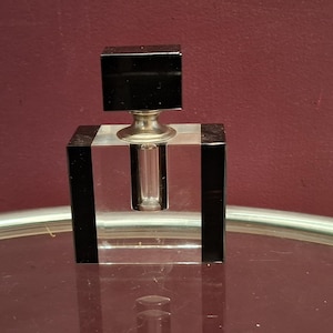 May include: A clear glass perfume bottle with a black square stopper. The bottle has a rectangular base with black vertical stripes. The stopper is also a black square. The bottle sits on a reflective surface.