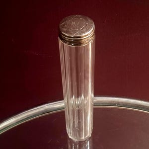 Peut inclure: Un grand récipient cylindrique en verre avec des détails cannelés verticaux et un couvercle circulaire en métal argenté. Le couvercle présente un motif floral gravé complexe sur sa surface supérieure. Le corps en verre est clair et transparent, affichant une forme élancée qui se rétrécit légèrement vers la base.