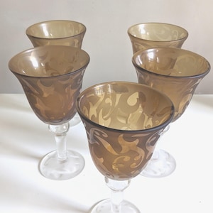 May include: Set of six amber-colored wine glasses. Each glass features a decorative etched design on the bowl. The glasses have a clear stem and base. The glasses are arranged on a white surface.