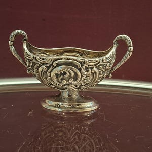 Pode incluir: Molheira prateada ornamentada com motivos florais e volutas. Possui duas alças curvas e uma pequena base redonda. O interior é dourado, contrastando com o exterior prateado. Uma peça decorativa.