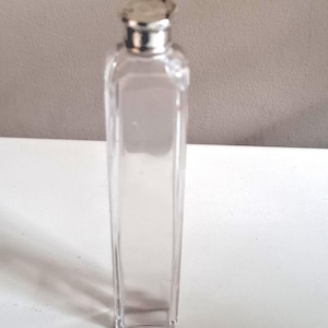 Puede incluir: Una botella de vidrio transparente y rectangular con una tapa plateada. La botella está vacía y tiene un diseño simple y elegante. La botella es probablemente para perfume u otros líquidos.