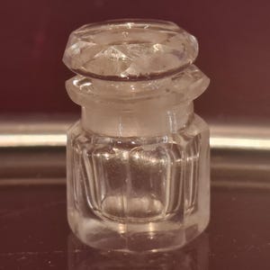 Pode incluir: Frasco de perfume pequeno, de vidro transparente, com tampa facetada. O frasco tem formato retangular com ranhuras verticais. A tampa também é facetada, adicionando um elemento decorativo.