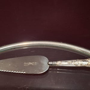 Pode incluir: Uma espátula de bolo prateada com borda serrilhada e cabo ornamentado. A lâmina é estampada com texto. A espátula repousa sobre uma superfície reflexiva, com borda prateada, contra um fundo escuro.