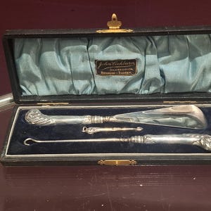 Könnte beinhalten: Ein Vintage-Silber-Pflegeset in einem schwarzen Etui mit blauem Satinfutter. Das Set enthält einen Schuhanzieher, einen Knopfhaken und ein gebogenes Werkzeug, alle mit verzierten Griffen. Das Etui ist mit "John Cockburn, Silversmith, Richmond - Surrey" beschriftet.