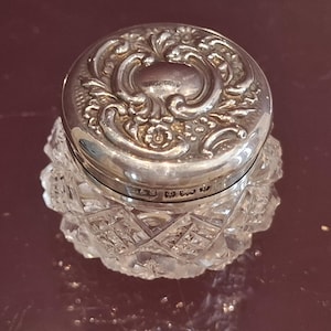 Puede incluir: Un pequeño tarro de cristal antiguo con tapa de plata. La tapa está decorada con diseños florales y de volutas. El cuerpo del tarro está tallado con un patrón de diamantes. Una pieza decorativa.