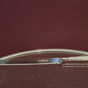 May include: A silver serrated knife with an ornate handle, resting on a reflective surface. The knife has the text "Algonquin Sterling Quality" etched on the blade. The background is a deep burgundy colour.