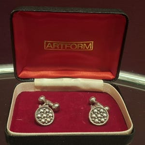 May include: A pair of silver cufflinks with a textured, circular design, presented in a black box with a red velvet interior. The box has the gold text "ARTFORM" on the inside of the lid. The cufflinks are a classic accessory.