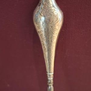 May include: A silver-toned metal utensil handle featuring an elongated, teardrop-shaped top section decorated with intricate, engraved scrolling floral patterns. The handle tapers down to a slender, cylindrical shaft with horizontal ring details.