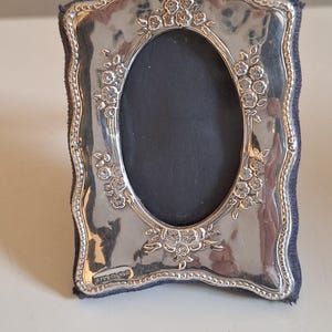 Puede incluir: Un marco de fotos ovalado de plata con borde festoneado y detalles florales en relieve. El marco presenta un respaldo de tela azul oscuro y una abertura ovalada central. Se observan pequeños sellos en la esquina inferior izquierda.