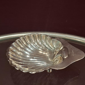 Op de afbeelding: Een zilverkleurige, schelpvormige schaal met een geribbelde textuur. De schaal heeft een kleine, platte basis en een decoratief, wervelend ontwerp aan één kant. De schaal is waarschijnlijk gemaakt van metaal en wordt gepresenteerd op een reflecterend oppervlak.