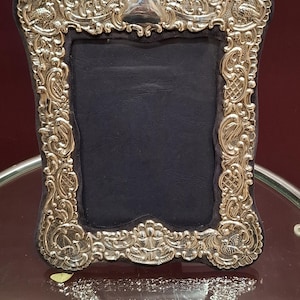 May include: Ornate silver-colored picture frame with a black interior. The frame features detailed scrollwork and decorative elements. The frame is rectangular with a curved top and bottom. The frame is sitting on a glass surface.