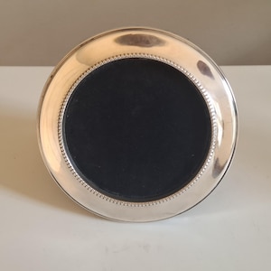May include: A circular silver-toned picture frame featuring a polished metallic border and a delicate beaded inner rim detail. The centre of the frame is filled with a solid black circular insert, providing a stark contrast to the reflective silver finish.