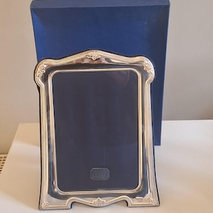 May include: A rectangular silver picture frame with ornate, curved edges and a polished metallic finish. The frame features a dark blue velvet backing and a small rectangular label at the bottom centre that reads Sterling Silver.
