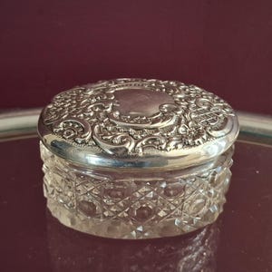 Könnte beinhalten: Ein rundes Glasgefäß mit einem klaren Diamantschliffmuster an den Seiten und einem schweren, polierten Deckel aus silberfarbenem Metall. Der Deckel zeigt ein kompliziertes, geprägtes Blumen- und Rankenmuster um ein glattes, rundes Medaillon.