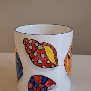 May include: White ceramic mug with a blue rim, decorated with colourful cartoon seashells and fish. The illustrations include red, orange, yellow, and blue designs. A fun, nautical-themed mug.