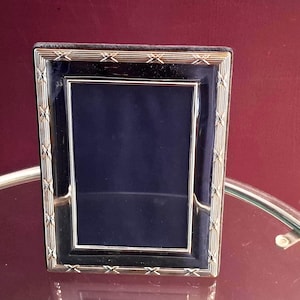 Op de afbeelding: Een rechthoekige fotolijst met een gepolijste zilverkleurige metalen rand met een herhalend X-vormig patroon. De lijst heeft een diep marineblauwe emaille inleg rondom de centrale opening die is ontworpen om een foto in te plaatsen.
