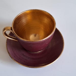 May include: A burgundy teacup and saucer set with gold trim. The interior of the cup is gold. The handle and rim of the saucer are also gold. The set is on a white surface.