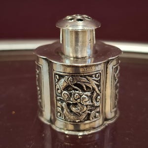 May include: A small, silver container with an elaborate design. The body of the container has detailed relief carvings of floral and mythical motifs. The container is likely an antique or vintage item.
