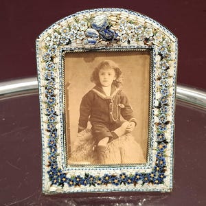 Könnte beinhalten: Ein antiker Fotorahmen mit einem floralen Mosaikdesign in Blau und Weiß. Der Rahmen umrahmt ein sepiafarbenes Porträt eines Kindes. Der Rahmen hat eine gewölbte Oberseite und steht auf einer reflektierenden Oberfläche.