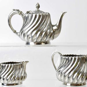 Può includere: Un servizio da tè in argento con teiera, lattiera e zuccheriera. Il set ha un design scanalato ed è esposto su una superficie trasparente. La teiera ha un coperchio con un pomello.