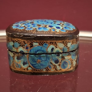 May include: A small, round, antique box with a hinged lid. The box is painted black with a floral design in shades of blue and gold. The flowers are clustered on the lid and around the sides.