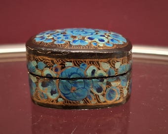 Può includere: Una piccola scatola rotonda e antica con coperchio a cerniera. La scatola è dipinta di nero con un motivo floreale nei toni del blu e dell'oro. I fiori sono raggruppati sul coperchio e sui lati.