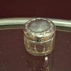 May include: A small, clear glass jar with a silver-colored lid. The lid has a decorative, scalloped edge. The jar sits on a reflective, dark surface, suggesting a vintage or antique item. The jar's sides are faceted.