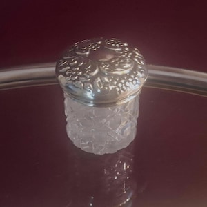 May include: A small, clear glass jar with a faceted, geometric cut pattern on its body. The jar is topped with a round, polished silver-toned metal lid featuring an embossed, intricate floral or leaf-like design across its entire surface.