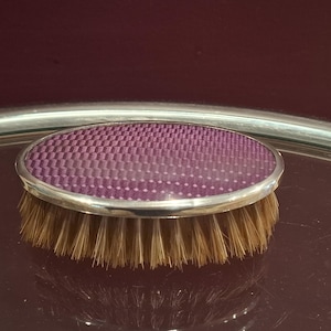 Op de afbeelding: Een ovale borstel met een zilverkleurige metalen rand en een bovenzijde met een paars patroon in koolstofvezelstijl. De basis van de borstel bestaat uit dichte, lichtbruine natuurlijke haren die in een gelijkmatige cirkelvorm zijn geplaatst.