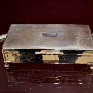 May include: A rectangular silver box with a textured lid and a small rectangular accent. The box has a hinged lid and rests on four small feet. The box is reflective, with a dark background.