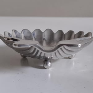 Pode incluir: Um pequeno prato prateado com borda ondulada e três pés esféricos. O prato tem um design em forma de concha e é provavelmente feito de metal. É um item decorativo.