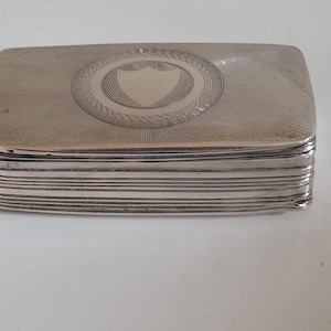 Könnte beinhalten: Eine rechteckige Silberbox mit Scharnierdeckel. Der Deckel zeigt ein zentrales Schilddesign, das von einem dekorativen Rand umgeben ist, mit radialen Linien, die sich zu den Rändern erstrecken. Die Box hat ein geschichtetes Seitendesign.