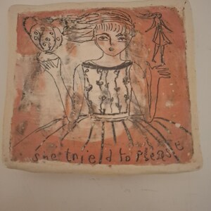 Puede incluir: Pieza de arte de cerámica cuadrada con una ilustración dibujada a mano de una niña sosteniendo un objeto decorativo y una figura más pequeña. El fondo es de color rojo anaranjado, y el texto "she tried to please" está escrito en la parte inferior.