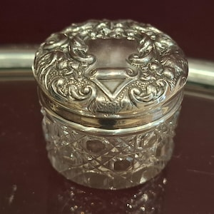 Puede incluir: Un pequeño frasco redondo de vidrio con un patrón de corte de diamante transparente en los lados. El frasco cuenta con una tapa extraíble de plata pulida decorada con intrincados patrones florales y de volutas en relieve alrededor de un área central lisa.