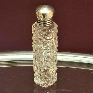 Könnte beinhalten: Eine kleine, klare Glasparfümflasche mit silberfarbenem Verschluss und Hals. Die Flasche hat ein rautenförmiges Muster, das das Licht reflektiert. Die Flasche steht auf einem silberfarbenen Tablett mit dunkelrotem Hintergrund.