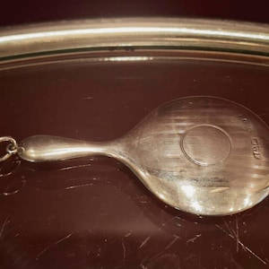 May include: A silver-toned metal charm shaped like a hand mirror with a round head and a slender handle. The surface features a circular centre detail and small stamped hallmarks on the right side. A small circular jump ring is attached to the end.
