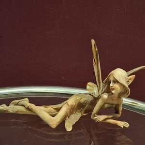 May include: A figurine of a fairy with blonde hair, pointed ears, and delicate wings. The fairy is lying down, with one arm bent and the other extended. The fairy is wearing a light brown dress. The background is a dark red color.