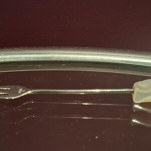 May include: A small, silver fork with three tines and a light-coloured, rectangular stone at the end of the handle. The fork is resting on a reflective surface, with a dark background.