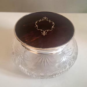 May include: A clear glass vanity jar with a rounded body featuring etched floral patterns. The jar has a circular lid with a dark brown tortoiseshell-like finish, a silver-toned metal rim, and a central silver filigree decorative emblem.