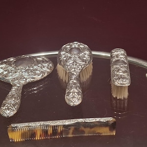 May include: A four-piece vanity set comprising a silver-toned hand mirror, a hairbrush, a clothes brush, and a tortoiseshell-patterned comb. Each item, with the exception of the comb, features an intricate, embossed floral and scrollwork metal design.