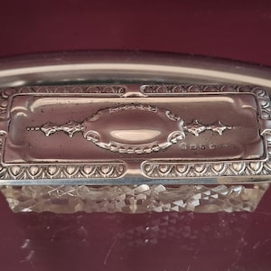 May include: A rectangular antique silver box with a faceted crystal base. The silver lid features an oval design and decorative accents. The box is displayed on a reflective surface, showcasing its intricate details and craftsmanship.