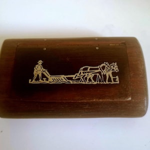 May include: A rectangular wooden box with a hinged lid featuring a metallic inlay of a person guiding a plough pulled by two horses. The box has a dark brown finish with visible wood grain and a smooth, rounded edge design on the top lid surface.