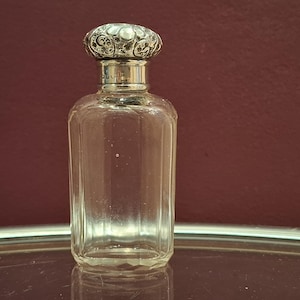 May include: A clear, rectangular glass perfume bottle with a decorative silver-colored lid. The lid features an ornate design and a small, round, pearl-like embellishment. The bottle is empty and sits on a reflective surface against a burgundy background.