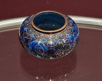 May include: A small, round, blue cloisonné vessel with a dark blue interior and a gold-coloured rim. The exterior features a floral pattern in shades of blue, black, and gold. The vessel is sitting on a reflective surface.