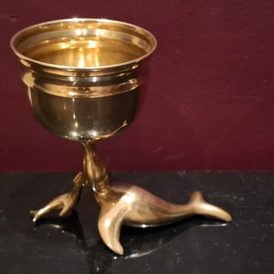 Könnte beinhalten: Ein Messingkelch mit einzigartigem Design. Die Tasse wird von einem Stiel getragen, der in eine stilisierte Seehundfigur übergeht. Der Kelch besteht aus poliertem Messing und steht auf einer schwarzen Oberfläche vor einem bordeauxroten Hintergrund.