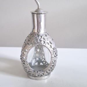 May include: A silver-coloured decorative oil bottle with a clear glass interior. The bottle has an ornate, openwork design around the glass, and a curved spout. The bottle sits on a white surface.