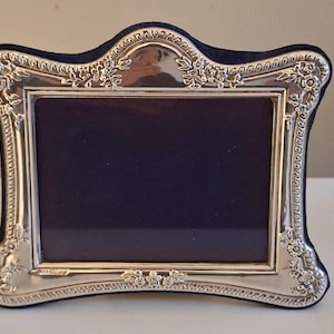May include: A silver-coloured picture frame with a dark blue interior. The frame features an ornate floral design around the edges and a dark blue velvet backing. The frame is rectangular with curved edges.