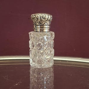 May include: A clear, cut-glass perfume bottle with a silver-coloured, ornate lid. The bottle has a diamond-like pattern and a silver band at the neck. The lid features a detailed, swirling design. The bottle is displayed on a reflective surface.