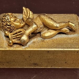 May include: A gold-colored, rectangular box with a textured surface, featuring a detailed, reclining cherub sculpture with wings. The cherub is holding a lyre, and the box has a vintage aesthetic.